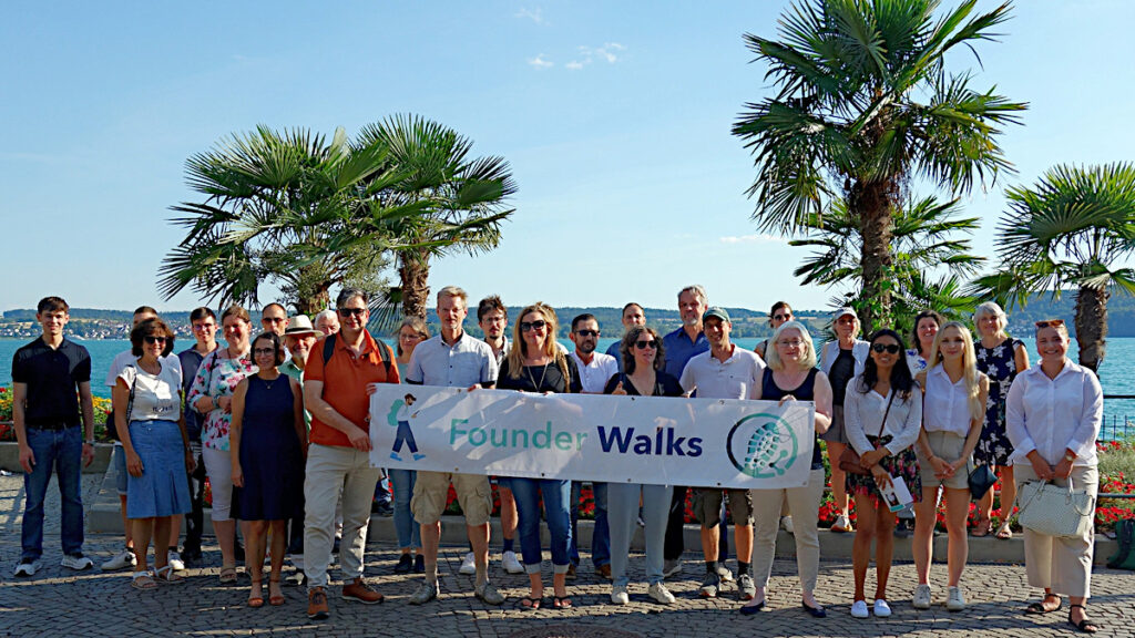 Gruppenbild Teilnehmende Founder Walks am 2. Juli 2026 in Überlingen am Bodensee. Die Gruppe hält ein "Founder Walks"-Banner im Vordergrund. Im Hintergrund sieht man Palmen und den Bodensee.
